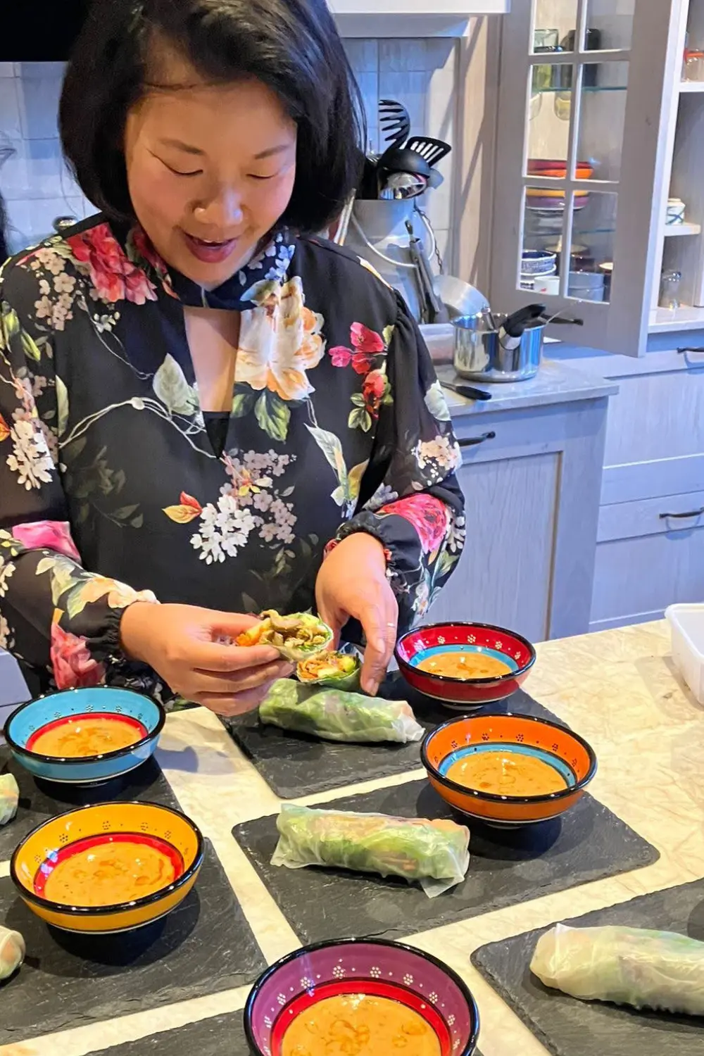 Nui joyfully plating fresh spring rolls with peanut sauce on 6 stone plates and colorful Tunisian bowls on a kitchen counter.