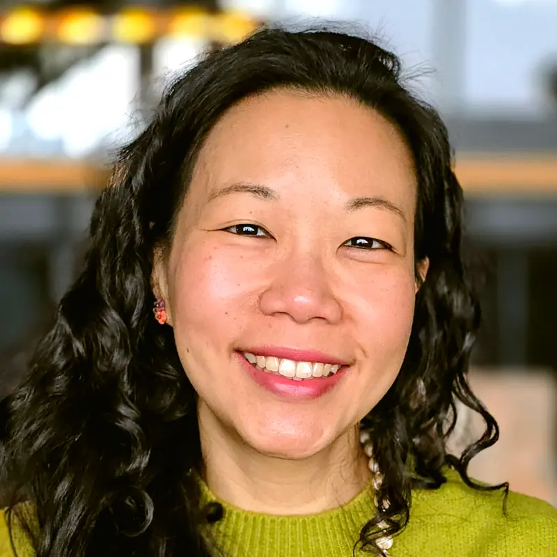 Close portrait of smiling Nui in a green sweater with a champagne bead necklace and peach earrings.