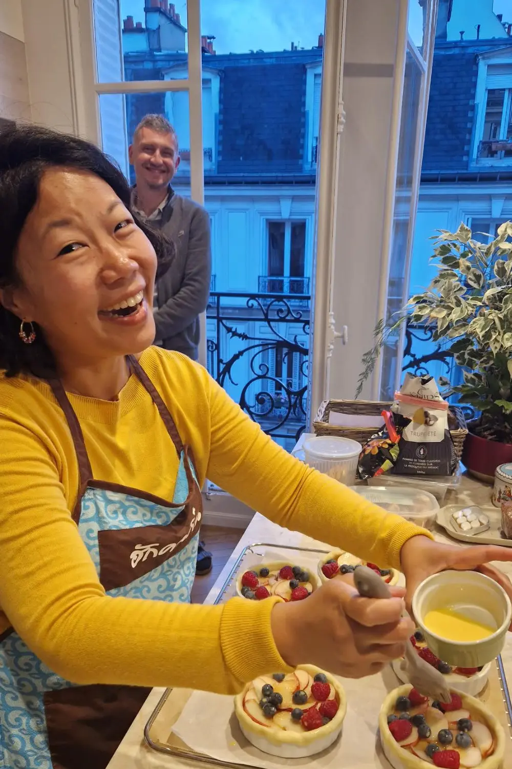 Smiling Nui in a yellow top and blue apron winks while making berry tarts, with Nicolas smiling in the background.