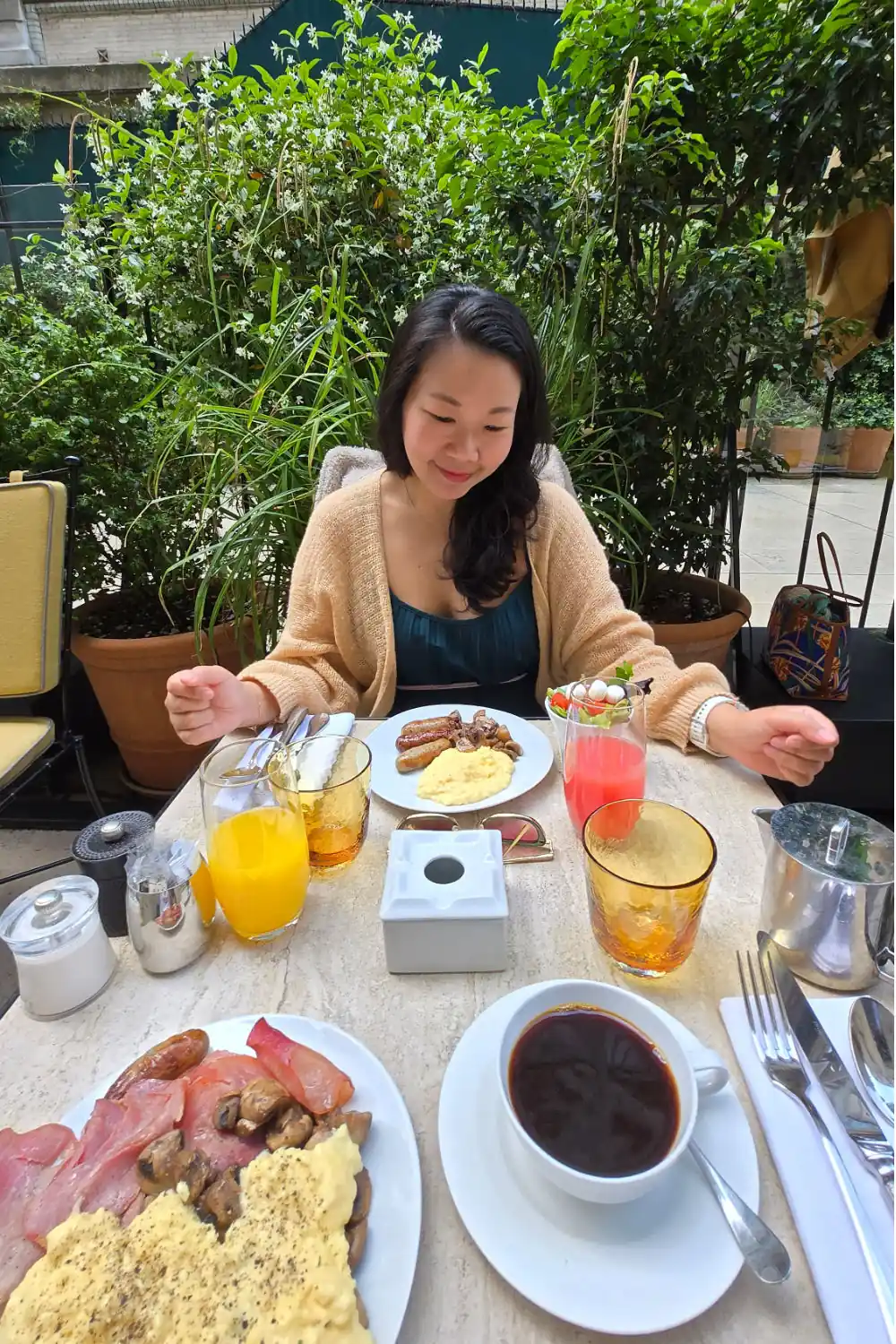Nui adoring a lavish breakfast spread with sausages, eggs, fresh fruit juices, and coffee outdoors on a leafy terrace.