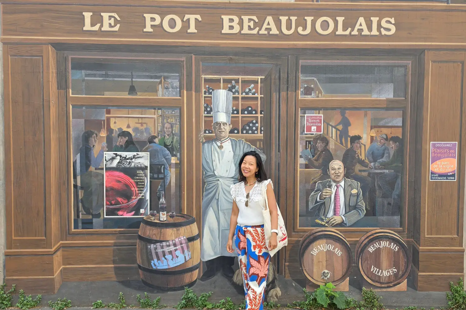 Smiling Nui at the Fresque des Lyonnais mural, featuring Paul Bocuse at "Le Pot Beaujolais" restaurant, with wine barrels.