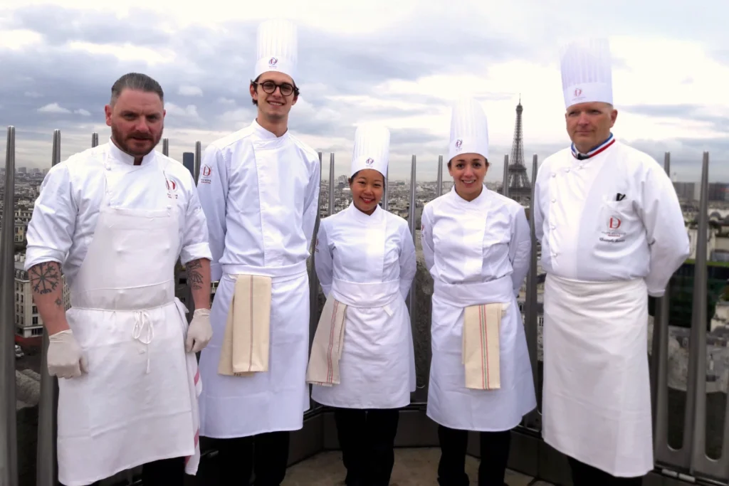Nui in École Ducasse chef uniform with instructors and classmates on the Arc de Triomphe rooftop with Eiffel Tower backdrop.