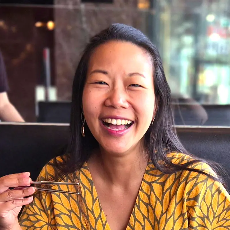 Nui at a restaurant table, wearing a yellow patterned top, holding chopsticks and smiling brightly.