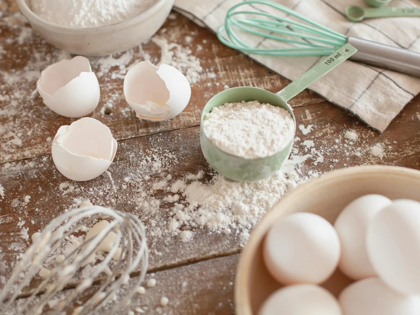 Baking scene: flour, eggs, whisks, and measuring cup on wood.