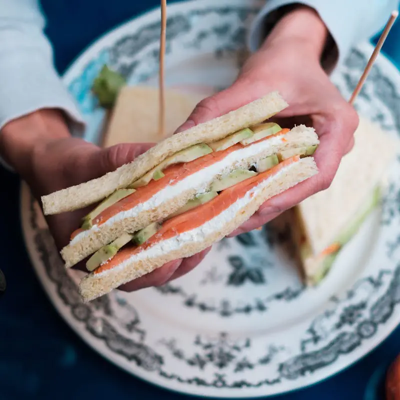 Salmon and avocado tea sandwich held over plate.