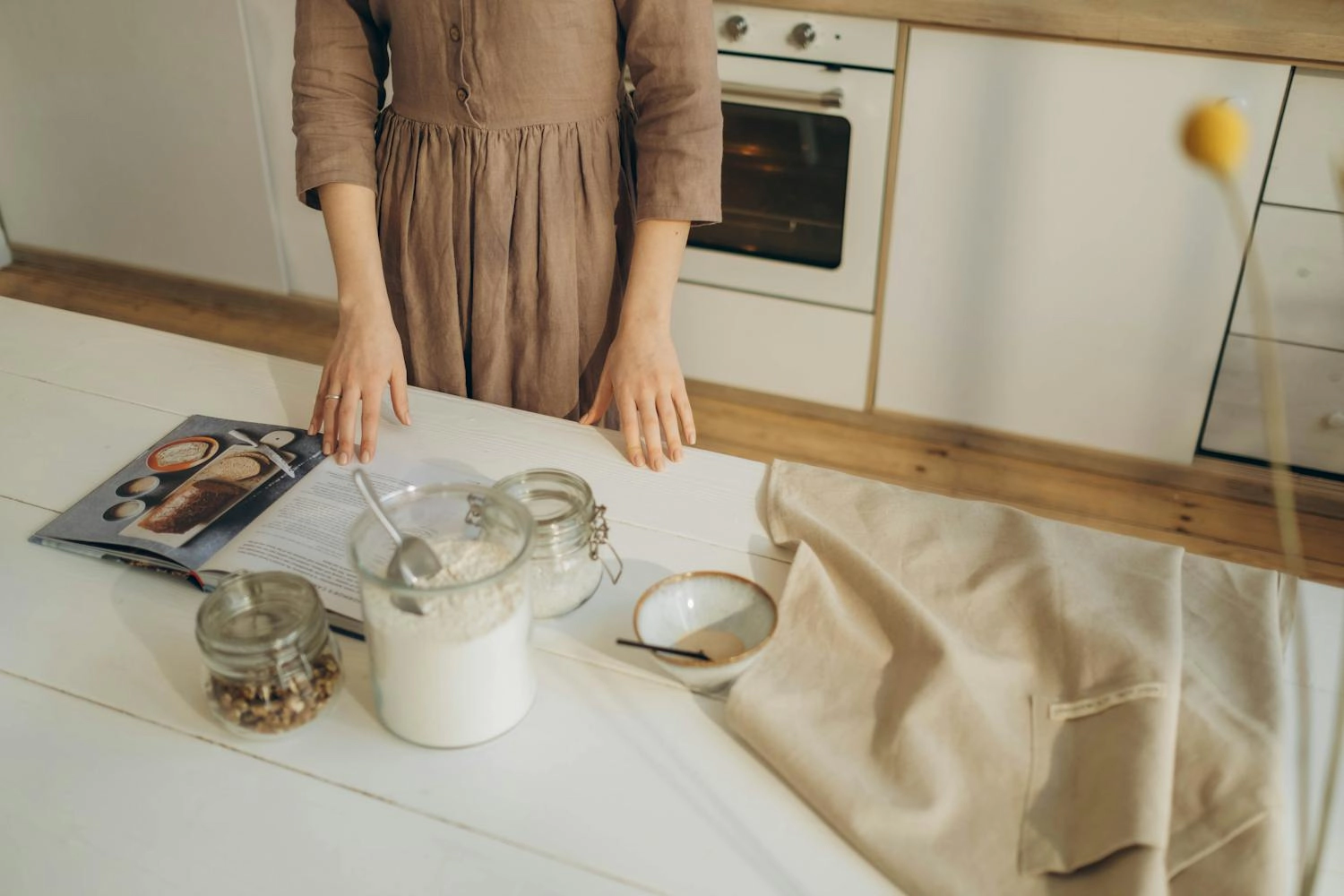 Woman baking in kitchen with cookbook, flour, and kitchen towel on counter.