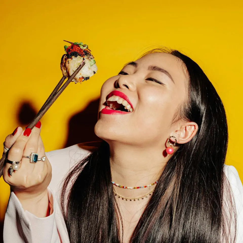 Woman joyfully eating sushi with chopsticks against a yellow background.